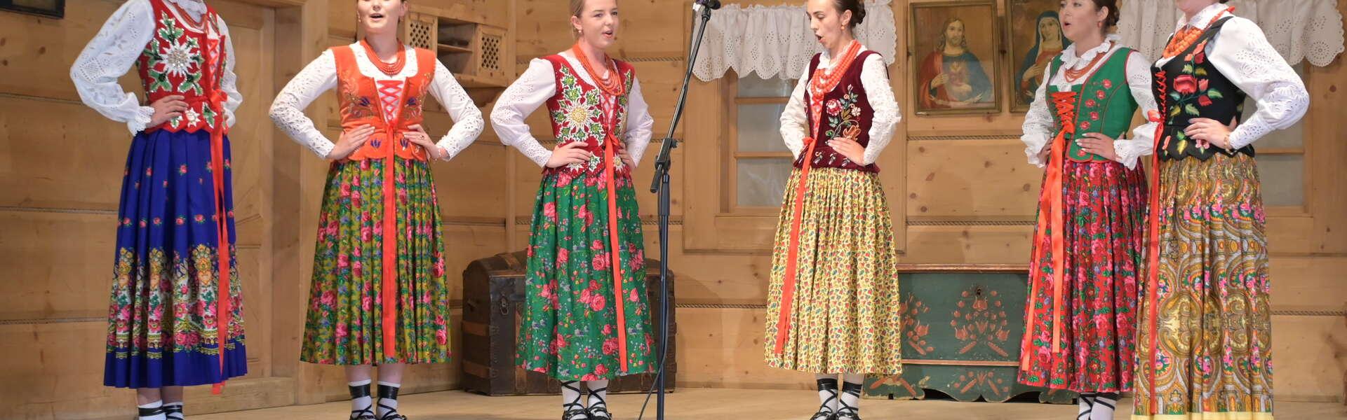 Sechs Frauen in regionaler Tracht, d.h. mit Hemden, Miedern und langen Röcken, stehen in einem Halbkreis auf einer Holzbühne. Vor ihnen ein Mikrofon, die Frauen singen und halten die Hände an ihren Hüften.