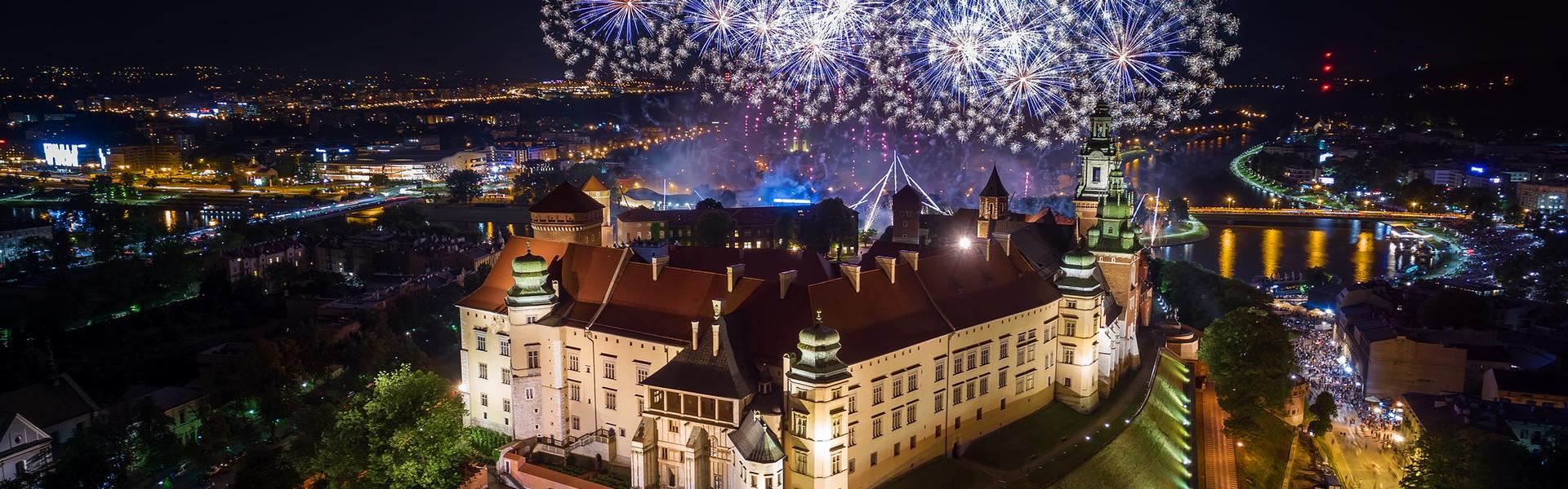 Oświetlone Wzgórze Wawelskie nocną porą od strony ul. Grodzkiej, nad nim sztuczne ognie wystrzelone znad Wisły