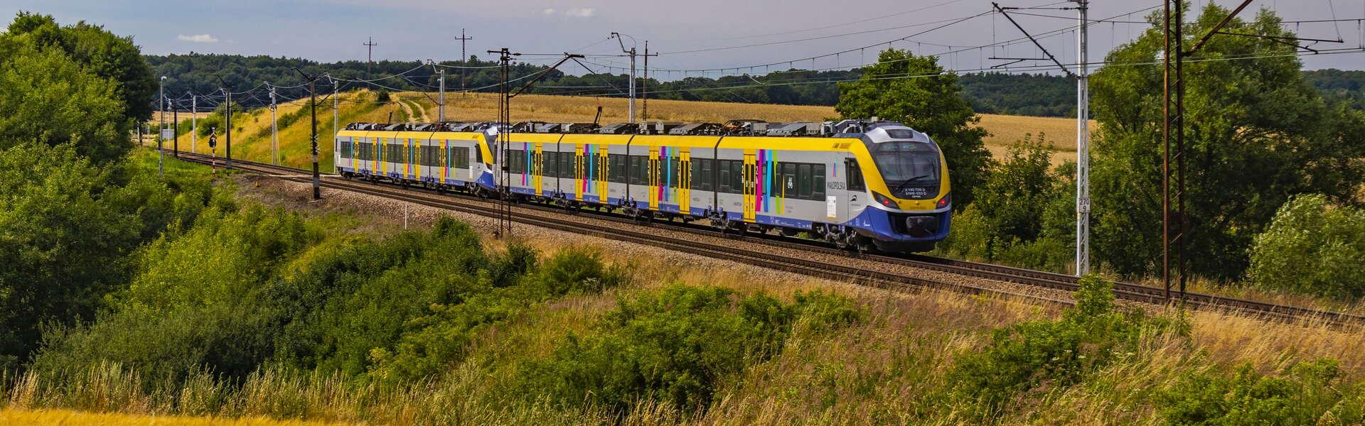 Widok na pociąg Kolei Małopolskich. Wokół pola i łąki.