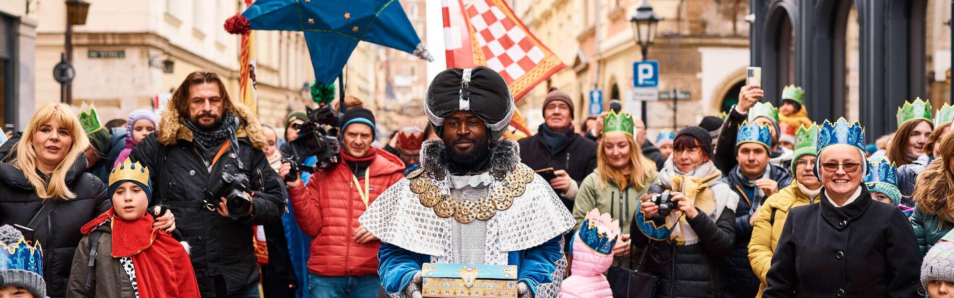 Na pierwszym planie mężczyzna przebrany za króla, w ręku trzyma drewnianą szkatułkę. W koło grupa osób, na głowach ma papierowe korony. Za królem osoba niesie gwiazdę betlejemską.